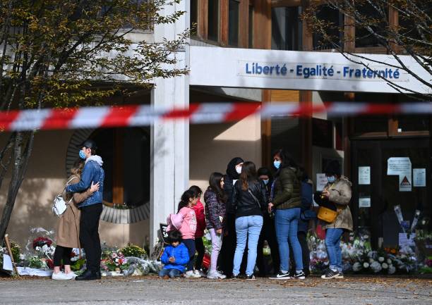 People gather near flowers layed outside the Bois d'Aulne secondary school in homage to slain history teacher Samuel Paty, who was beheaded by an attacker for showing pupils cartoons of the Prophet Mohammed in his civics class, on October 19, 2020, in Conflans-Sainte-Honorine, northwest of Paris. - Paty, 47, was attacked on October 16 on his way home from the junior high school where he taught by 18-year-old Chechen man Abdullakh Anzorov, who was shot dead by police. Following the attack, tens of thousands of people took part in rallies countrywide to honour Paty and defend freedom of expression. (Photo by Anne-Christine POUJOULAT / AFP) (Photo by ANNE-CHRISTINE POUJOULAT/AFP via Getty Images) | Epoch Times Россия