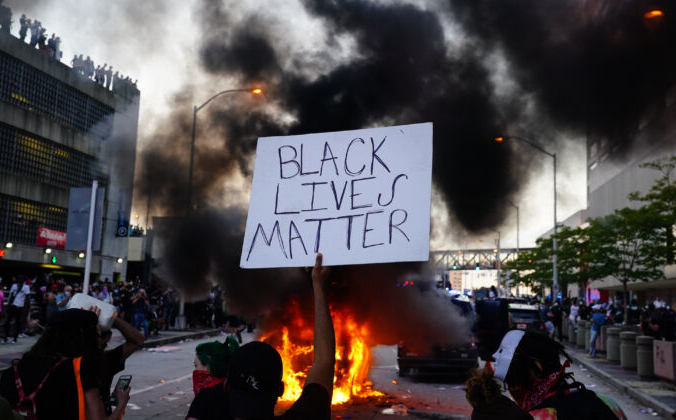 Мужчина держит знак Black Lives Matter, когда горит полицейская машина во время акции протеста в Атланте, Джорджия, 29 мая 2020 года. Elijah Nouvelage/Getty Images | Epoch Times Россия