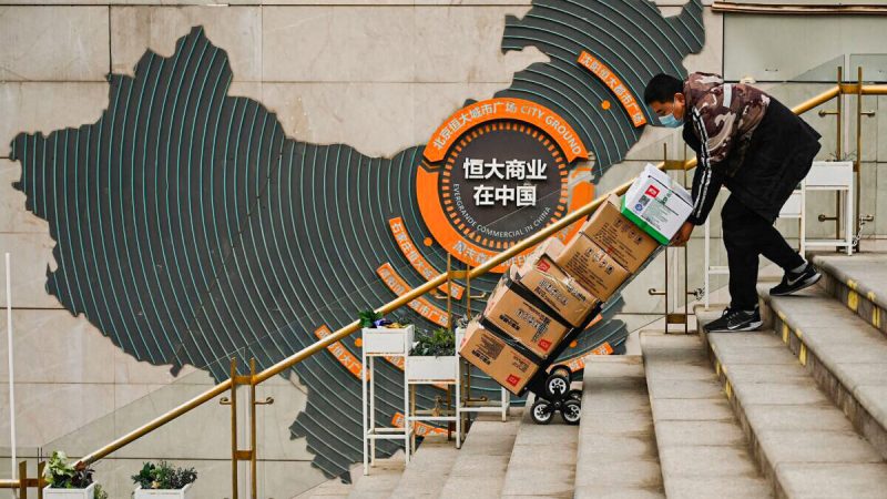 Рабочий толкает тележку перед вывеской, показывающей китайское подразделение Evergrande Group, в жилом комплексе застройщика в Пекине 8 декабря 2021 года. (Noel Celis/AFP via Getty Images)  | Epoch Times Россия