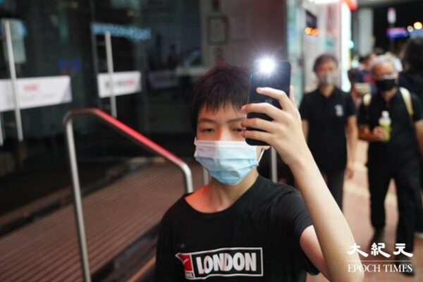 Подросток идёт по улице в Козуэй-Бэй, подняв над головой телефон с зажжённым фонариком. (Yu Gang/The Epoch Times, HK)