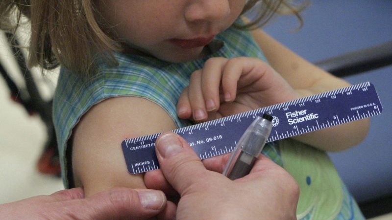 Ребёнок смотрит, как медсестра измеряет след, оставшийся на руке после прививки. (AP Photo/John Amis)  | Epoch Times Россия
