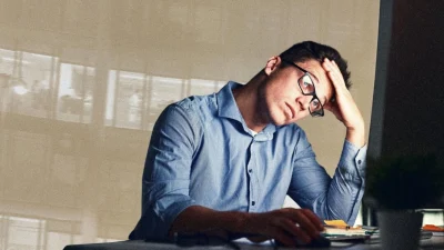 Уставший офисный сотрудник в очках сидит за компьютером, схватившись за голову от переутомления и стресса