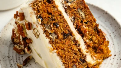 Кусочек морковного торта с кремом и посыпкой из орехов пекан на тарелке.