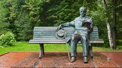 Бронзовая скульптура сидящего на скамье композитора Чайковского в Клину на фоне зеленых деревьев.