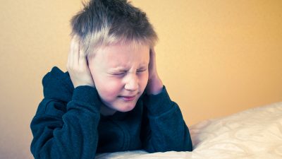 Мальчик с зажмуренными глазами плотно закрывает уши руками, демонстрируя нежелание слушать критику или крики.