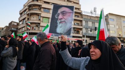 Толпа людей в Тегеране на траурном митинге; женщина на переднем плане держит большой плакат с портретом аятоллы Али Хаменеи на фоне городских зданий и флагов Ирана.