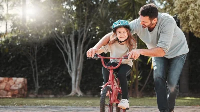 Заботливый отец помогает маленькой дочери учиться кататься на велосипеде в солнечный день, демонстрируя качественное времяпровождение с ребенком.