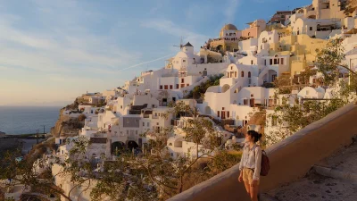 Девушка-туристка с рюкзаком смотрит на белоснежный город, расположенный на крутой скале у моря в лучах заката.