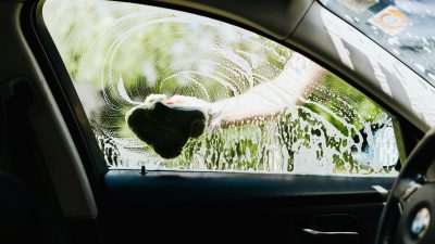 Процесс мойки стекла автомобиля губкой с пеной на фоне дачного участка и зелени.