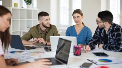 Группа молодых людей, студентов и специалистов, обсуждает рабочие задачи и графики за столом с ноутбуками в современном офисе